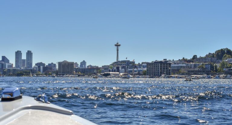 A view from a boat on sparkling blue water, looking toward the Seattle skyline with the Space Needle and tall buildings under a clear, sunny sky.