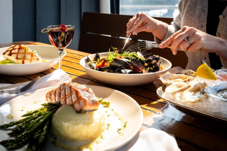 A table set with grilled fish on mashed potatoes and asparagus, a bowl of mussels, oysters on ice with lemon, and a glass of red wine. A person is using utensils to eat from the bowl of mussels.
