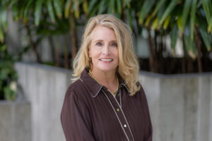 A woman with long blonde hair wearing a brown blouse with white piping and gold hoop earrings stands smiling in front of green leafy plants as Seattle names VP of meetings and conventions.
