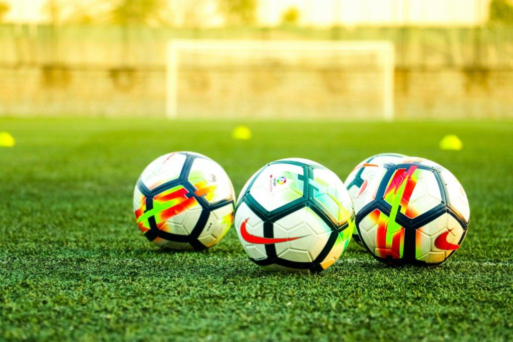 Three soccer balls with colorful patterns lie on green artificial turf, with a blurred goalpost and yellow training cones in the background, suggesting a practice or training session.