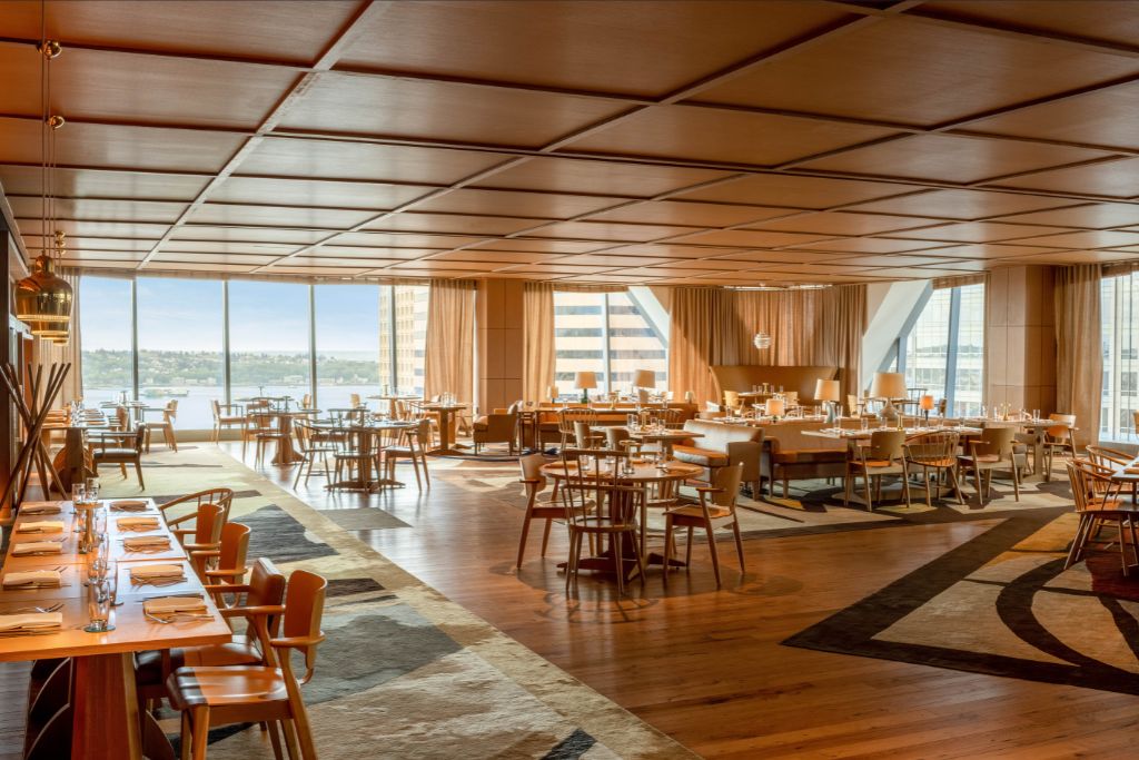A spacious, modern restaurant with wooden floors and ceiling, large windows with city and river views, and neatly arranged tables set for dining, creating a warm and inviting atmosphere.
