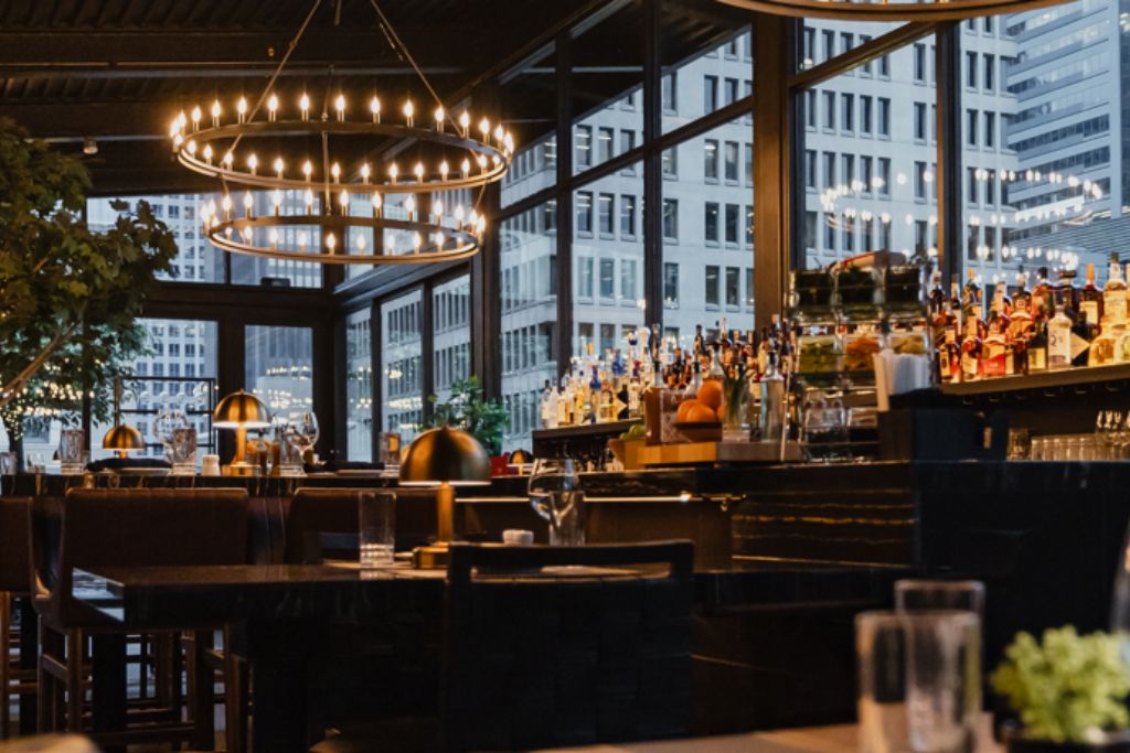 A modern, elegant restaurant interior with large windows showing city buildings, two circular chandeliers, a bar stocked with bottles, and neatly set tables with glasses and cutlery.