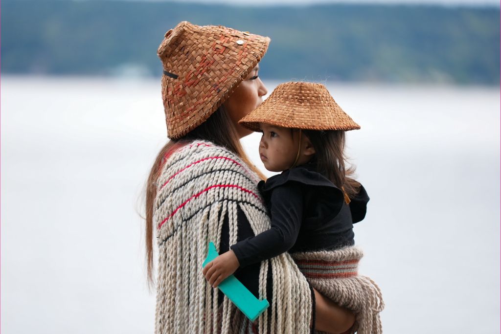 A woman wearing a woven hat and traditional attire holds a young child in similar dress. They stand outdoors with water and forested hills in the background. The child holds a small green object.