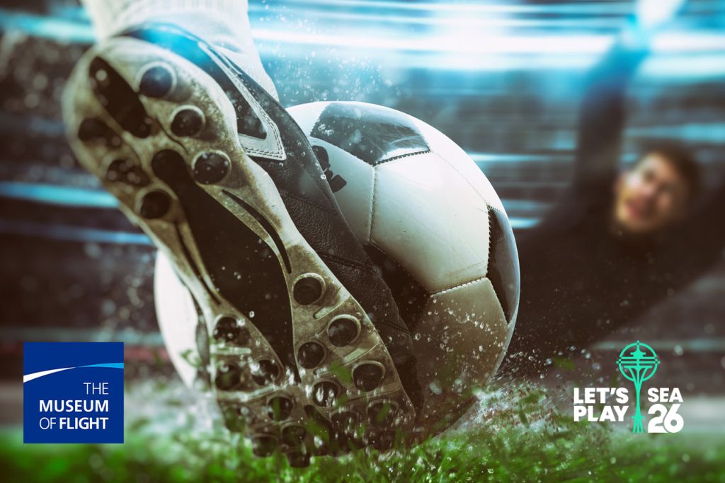 A close-up of a soccer player’s cleats kicking a wet soccer ball toward a diving goalie. Logos for The Museum of Flight and "Let’s Play SEA 26" appear in the lower left and right corners.