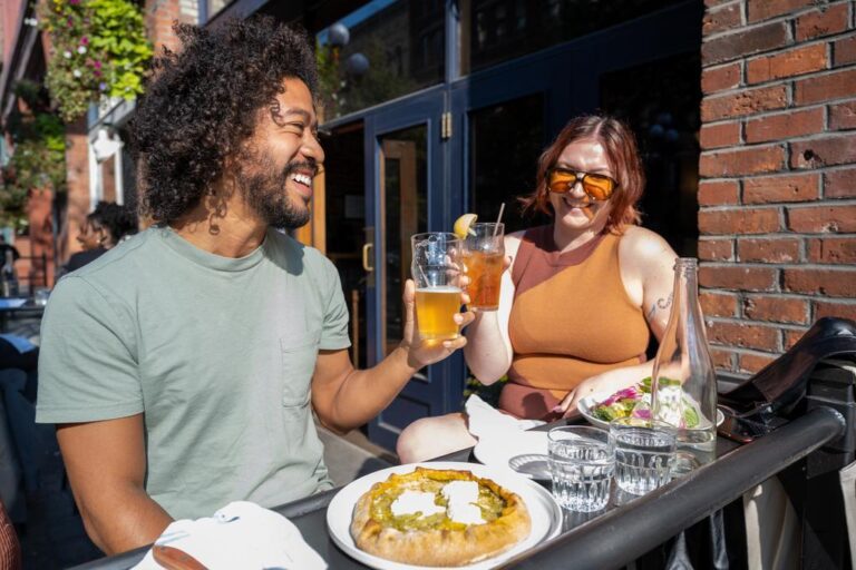 Two people sit at an outdoor restaurant table, smiling and toasting drinks. One has curly hair and wears a green shirt, the other has reddish hair, sunglasses, and an orange dress. Food and water glasses are on the table.