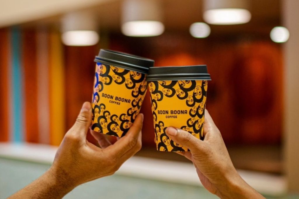 Two hands holding and clinking takeaway coffee cups with a yellow and black smiley face pattern and the text "BOON BOON COFFEE" against a blurred indoor background.