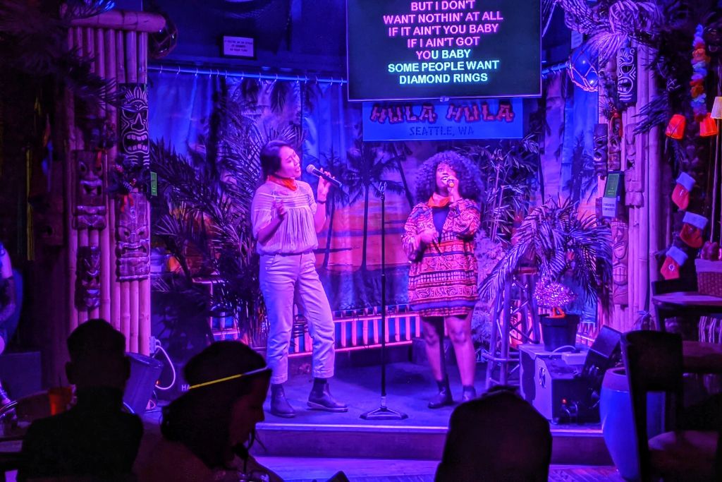 Two women sing karaoke on a colorful, tropical-themed stage with a screen above showing song lyrics. The background has palm decorations and tiki designs, and an audience watches from dimly lit tables.