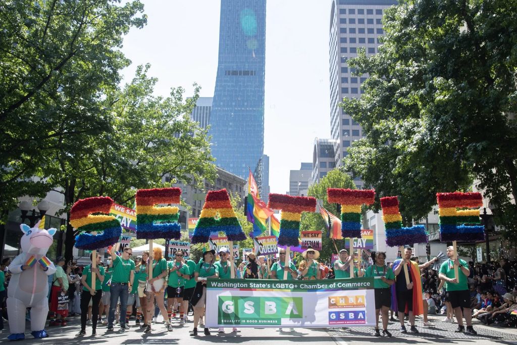 A group marches in a Seattle Pride parade holding large rainbow-colored letters spelling "SEATTLE" with banners from the LGBTQ+ Chamber of Commerce, surrounded by people in green shirts and festive costumes, under sunny skies and tall buildings.