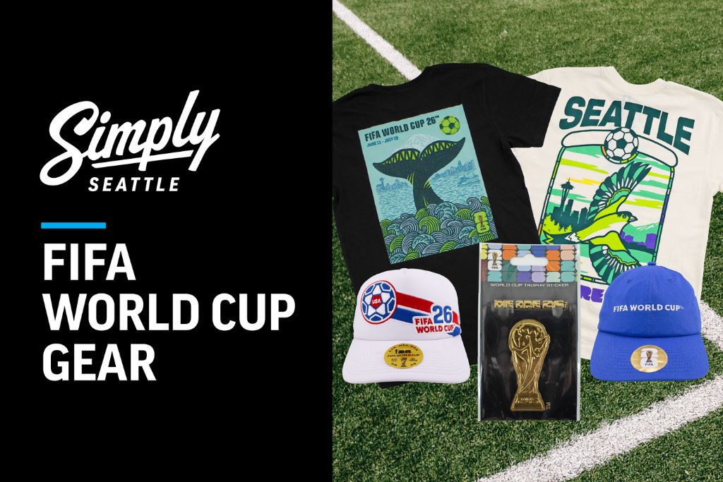 A display of Seattle FIFA World Cup gear on grass, including two graphic t-shirts, two hats, and a souvenir with a gold trophy image, beside a "Simply Seattle FIFA World Cup Gear" text on a black background.
