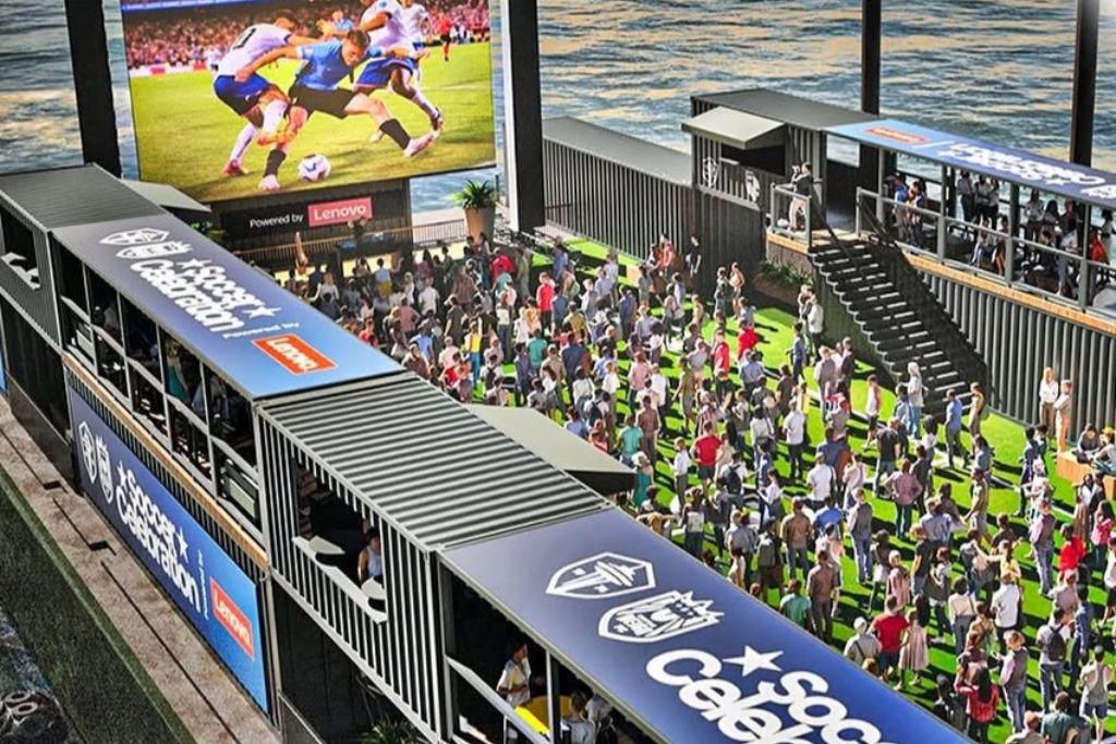 A large crowd gathers on a floating platform made of shipping containers, watching a soccer match on a big screen. The area is decorated with soccer banners and artificial grass, with water visible around the platform.