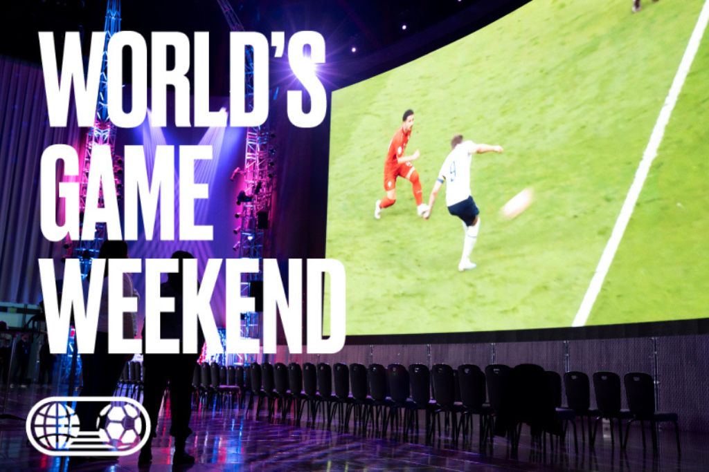 A large screen shows a soccer match with two players in action. The words "WORLD'S GAME WEEKEND" are in bold white letters, and empty chairs are lined up below the screen in a dark event space.