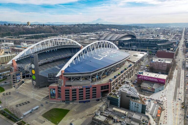 Aerial view of Lumen Field stadium in Seattle, with its arched roof and open seating. Nearby is T-Mobile Park, visible in the background, and city buildings and streets surrounding both venues.