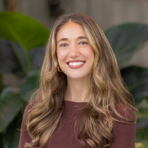 A woman with long, wavy light brown hair, wearing a maroon top and gold hoop earrings, smiles in front of large green leafy plants.