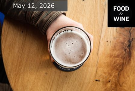 A person’s hand holding a frothy drink in a glass, viewed from above on a round wooden table. Text reads "May 12, 2026" in the top left and "FOOD & WINE" in the top right corner.