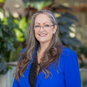 A woman with long, wavy gray hair and glasses smiles while wearing a royal blue blazer over a black top, standing in front of lush green plants in a bright indoor setting.