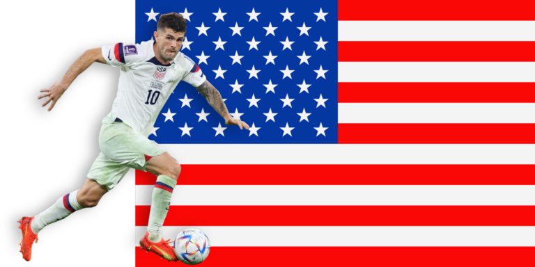A soccer player in a white uniform runs with a ball in front of a large United States flag, appearing focused and in action.