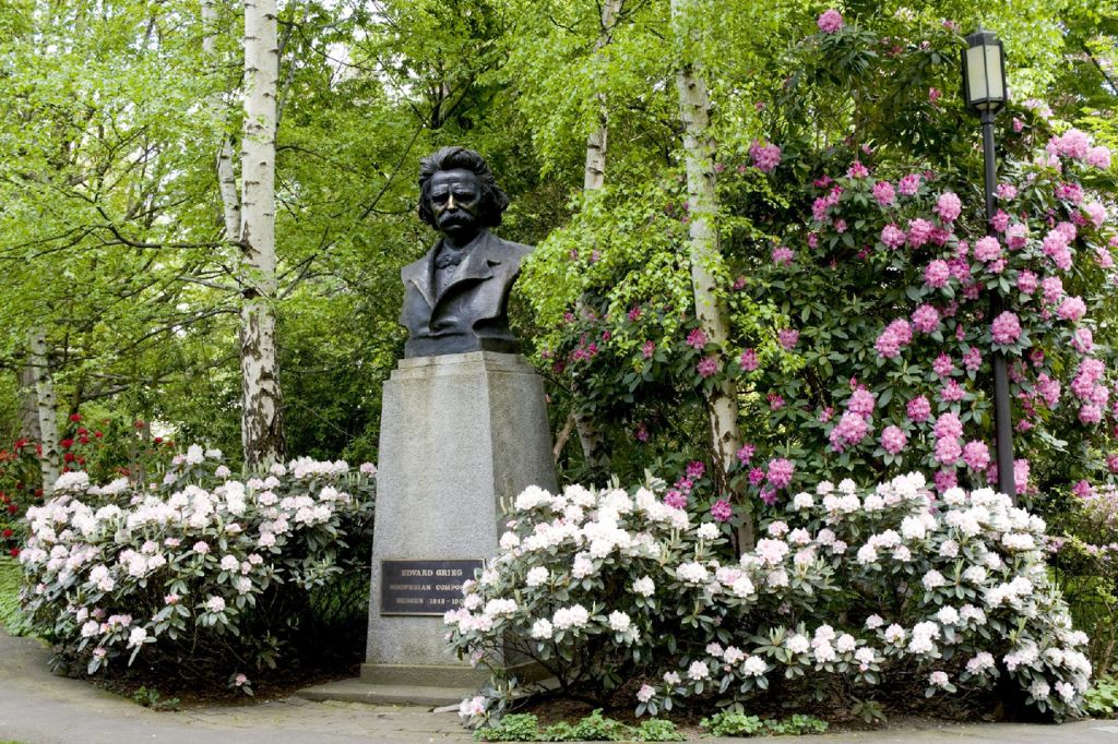 A bronze bust on a stone pedestal stands surrounded by blooming pink and white rhododendrons and green trees in a lush garden setting.