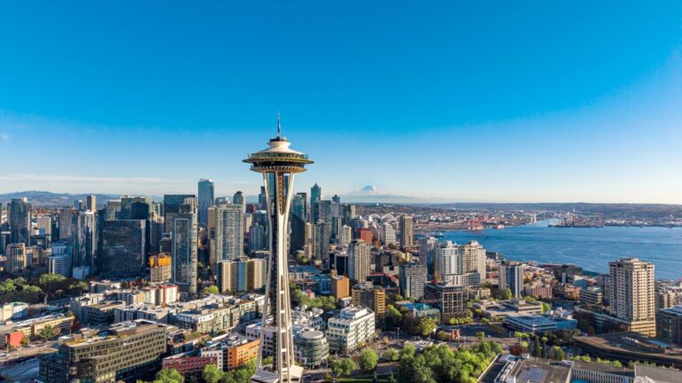 Seattle cityscape featuring the Space Needle in the foreground, downtown skyscrapers, Elliott Bay, and Mount Rainier in the distance under a clear blue sky.