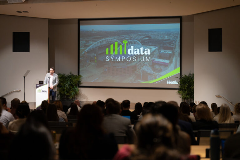 A speaker stands at a podium presenting to an audience at Data Symposium, with a large screen behind displaying the event's name and a city background.