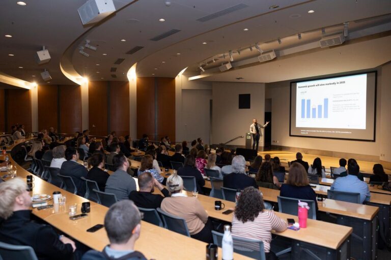 A speaker presents a bar graph on a large screen to an audience seated in a modern lecture hall. People are attentively watching the presentation, with notebooks, mugs, and water bottles on tables.