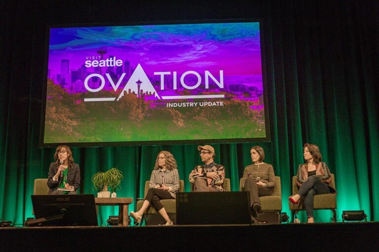Five women sit on stage in front of a large screen reading "Visit Seattle Ovation Industry Update" with a Seattle skyline graphic. One woman speaks into a microphone while the others listen.