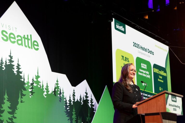 A woman stands at a podium speaking at the Visit Seattle Annual Meeting. Behind her, a screen displays 2025 hotel data and a backdrop features green trees and the Visit Seattle logo.