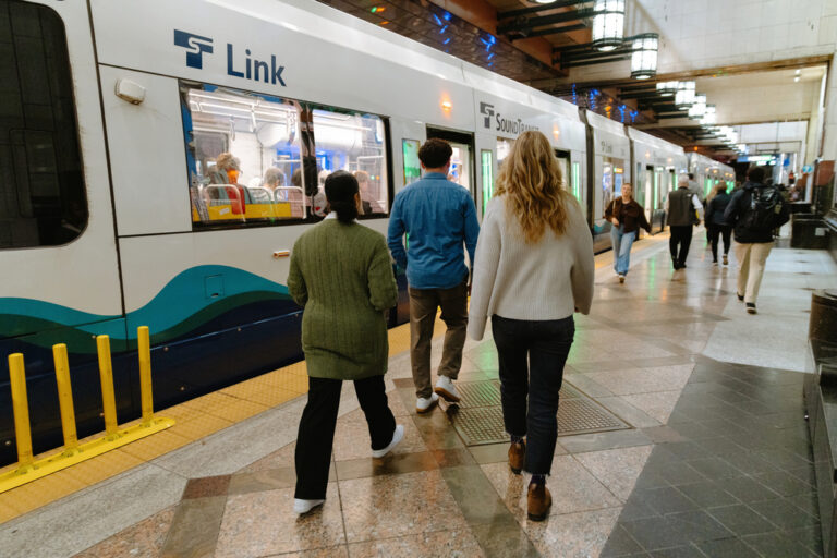 Three people walk toward a Link light rail train stopped inside a well-lit station, with several other passengers nearby and art deco-style lights overhead.