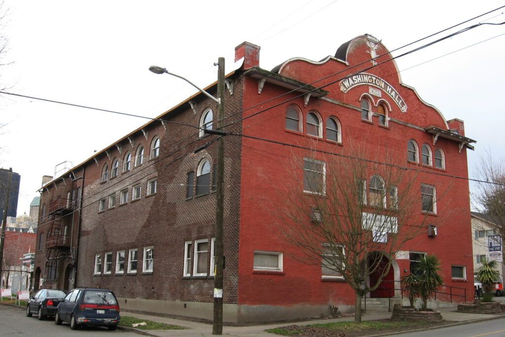 A large, historic red brick building with arched windows and a sign reading "Washington Hall" above the entrance, located on a street corner with parked cars and leafless trees.