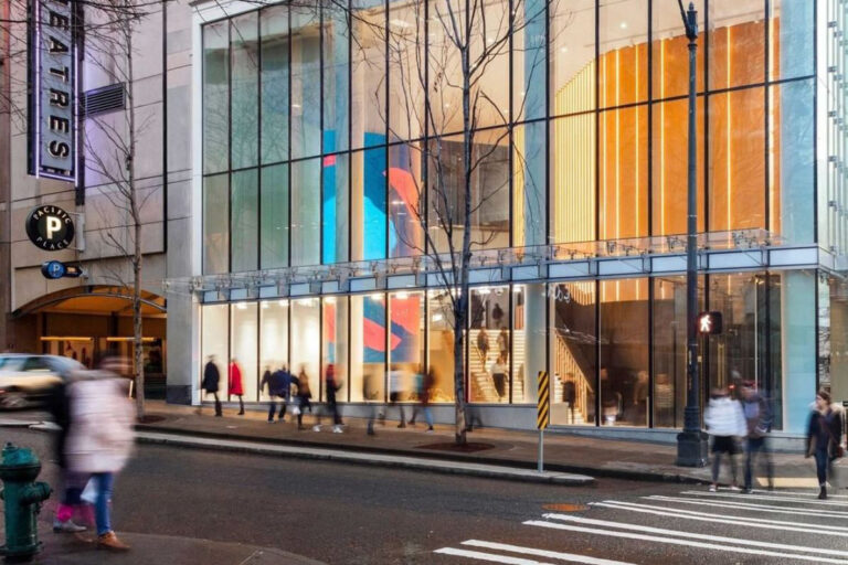 Street view of a modern, glass-fronted building with colorful interior art. People walk on the sidewalk, some blurred in motion. A crosswalk and traffic light are visible, along with a theater sign on the left.
