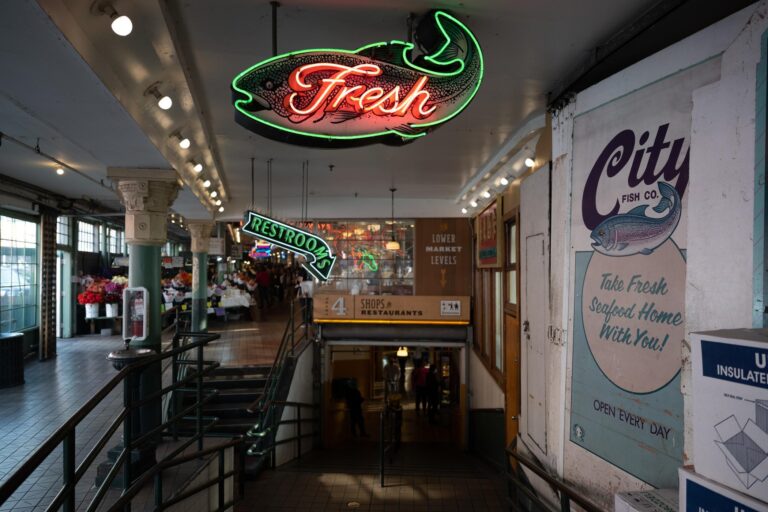 Neon signs for "Fresh" and "Restrooms" hang above a stairway leading to the lower level of an indoor market, with various posters and market stalls visible in the background.