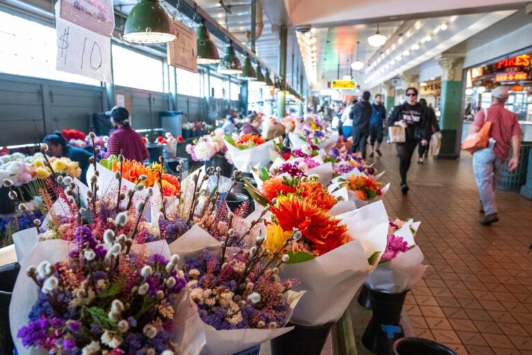 A vibrant indoor market scene with colorful bouquets of flowers displayed on tables, people walking and shopping, and overhead lights illuminating the bustling atmosphere.
