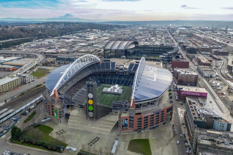 Aerial view of Lumen Field stadium in Seattle, surrounded by city buildings, roads, and railway tracks, with T-Mobile Park and Mount Rainier visible in the background under a cloudy sky.