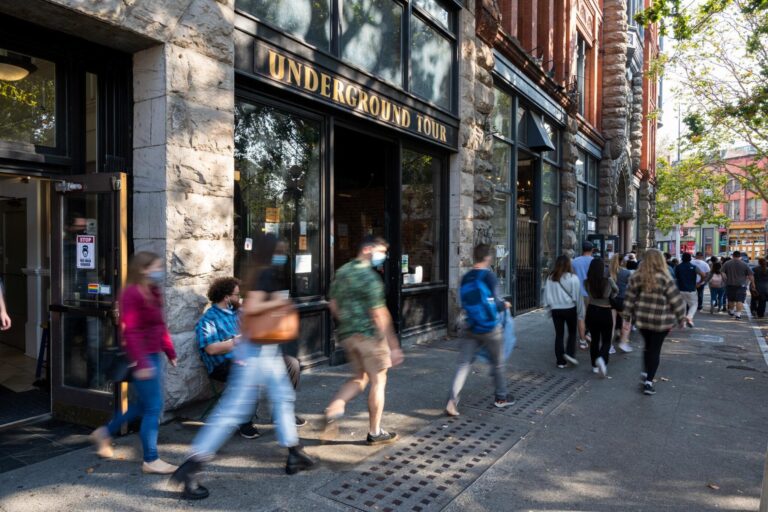 People walk along a city sidewalk past a building with a sign that reads "Underground Tour." Some individuals are sitting near the entrance, and the scene is lively with blurred motion from pedestrians.