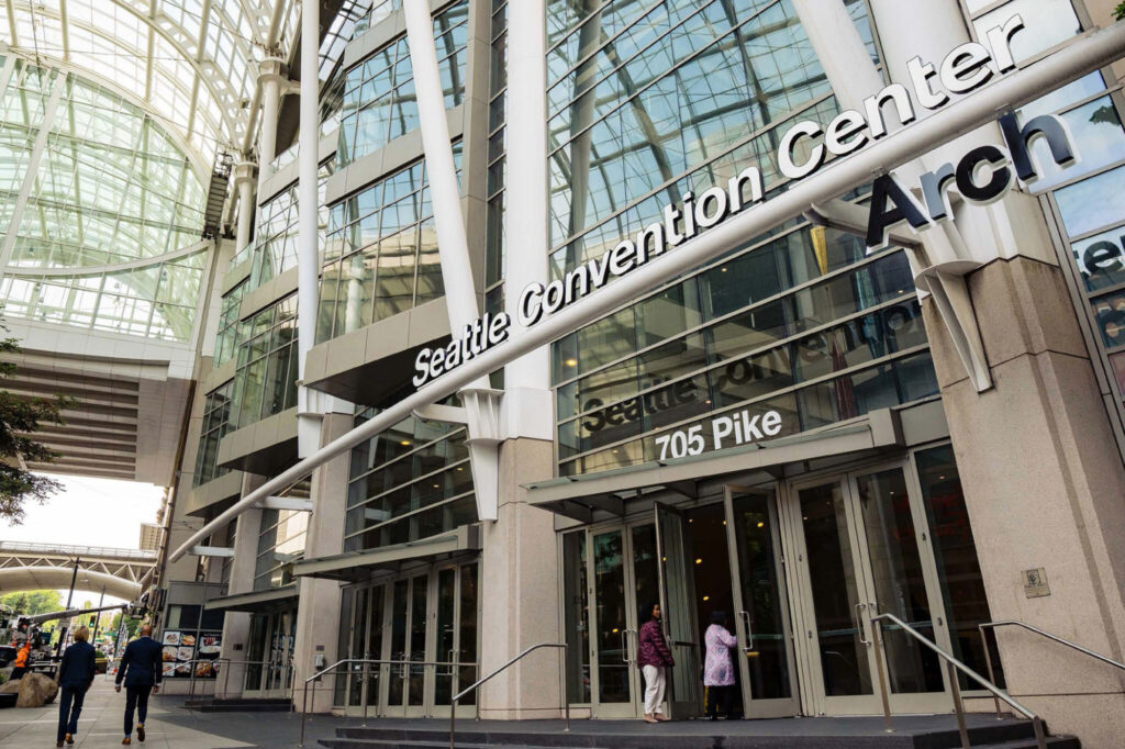 The entrance of the Seattle Convention Center Arch building at 705 Pike Street, with glass walls and people walking near the doors. The structure features modern architecture with tall glass and metal elements.