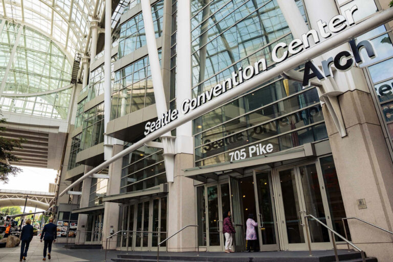 The entrance of the Seattle Convention Center Arch building at 705 Pike Street, with glass walls and people walking near the doors. The structure features modern architecture with tall glass and metal elements.