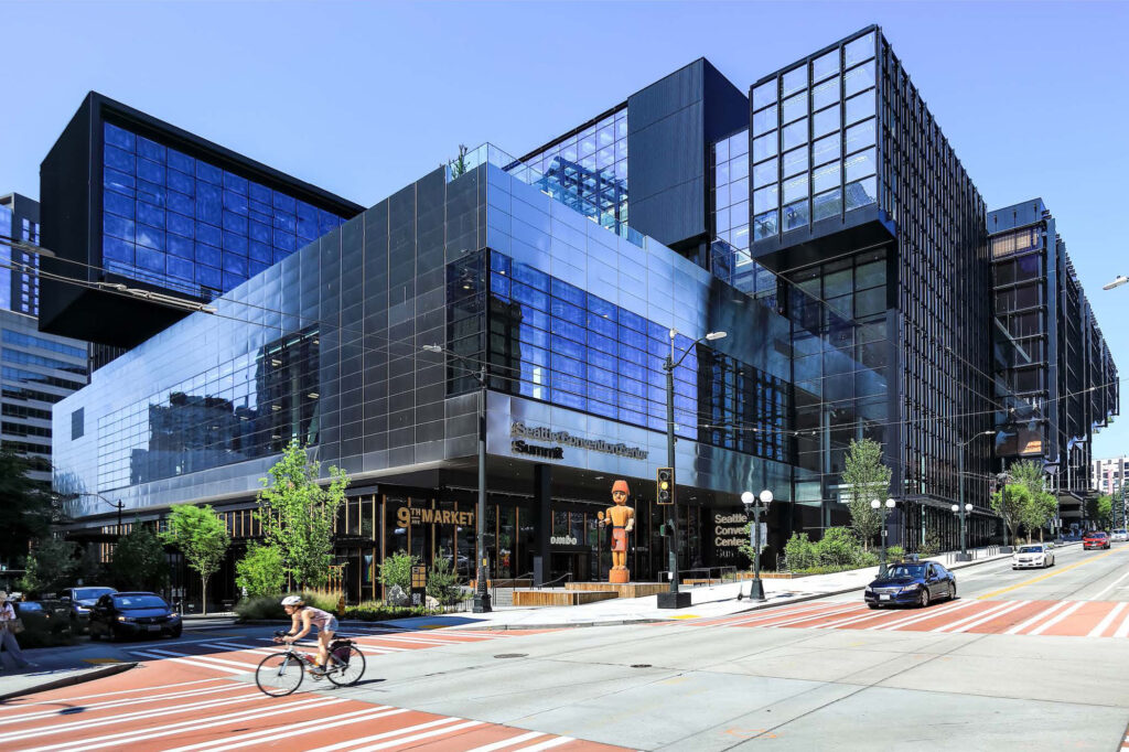 A modern glass building on a city corner houses the Seattle Convention Center Summit. A cyclist and a pedestrian cross the street, and cars are waiting at the traffic light under clear blue skies.