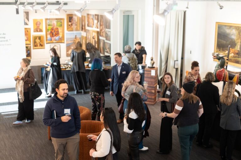 A group of people mingle and converse in an art gallery space, with paintings and sculptures on display. Several people are holding drinks and snacks, and some are viewing the artwork.