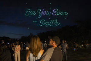 A couple stands together at night, looking up as drones form “See You Soon – Seattle” in glowing lights, celebrating as Seattle names VP of meetings and conventions—others watch this impressive sky display.