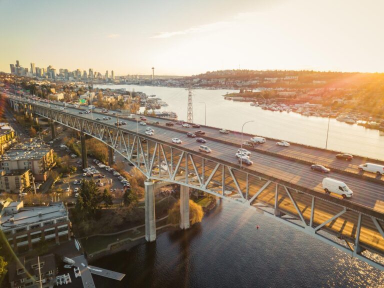 Aerial view of a busy bridge with cars and trucks crossing over water at sunset; a city skyline and the Space Needle are visible in the background.