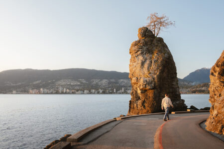A person walks along a curved path by the ocean, passing a large rock formation topped with a small tree. City buildings and mountains are visible across the water under a clear sky.