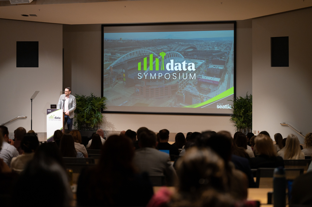 A man stands at a podium giving a presentation to an audience at the “Data Symposium.” The event’s logo and a cityscape image are displayed on a large screen behind him.