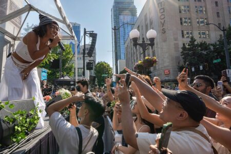 A singer performs energetically on an outdoor stage while a cheering crowd, some holding phones, gathers in front of a Macy’s storefront on a sunny day in the city.