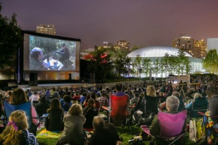 A large crowd sits on the grass at night, watching a movie projected on an outdoor screen in a city park, with buildings and trees illuminated in the background.