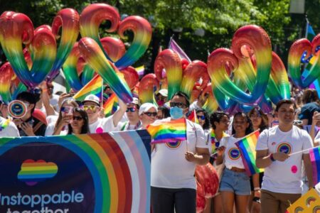 A crowd marches in a Pride parade, holding rainbow flags and heart-shaped rainbow balloons. Many people wear white shirts with rainbow designs, and trees are visible in the background. A colorful banner reads 