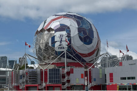 A large geodesic dome decorated like a soccer ball with the FIFA logo, surrounded by red, white, and grey buildings and several Canadian flags, under a partly cloudy sky.