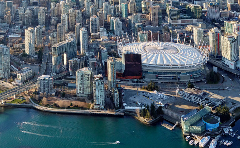 Aerial view of a cityscape featuring a large, circular stadium with a white roof, surrounded by high-rise buildings, roads, and waterfront with boats on blue water in the foreground.
