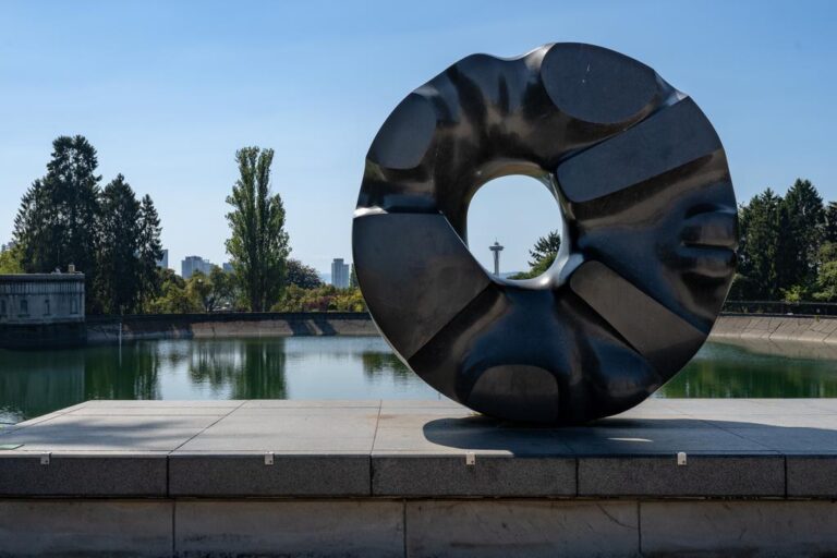 A large black abstract sculpture with a circular hole in the center sits on a concrete platform by a reflective pond, with trees and a distant city skyline, including the Space Needle, in the background.