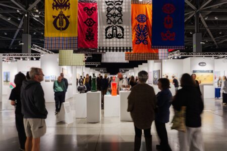 A crowd of people walk and observe artwork displayed on white stands in a brightly lit indoor art exhibition. Colorful patterned banners hang from the ceiling above the exhibits.