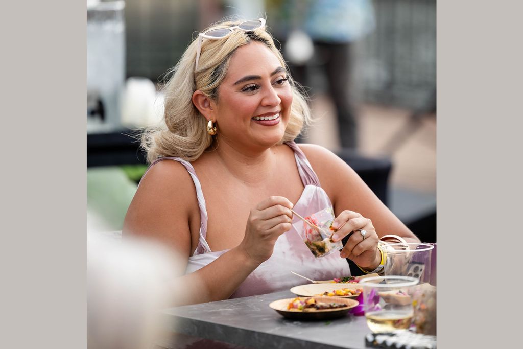 A woman with blonde hair and gold earrings smiles while dining outdoors, holding a small plastic bag of food. Plates of colorful dishes and a drink are on the table in front of her.