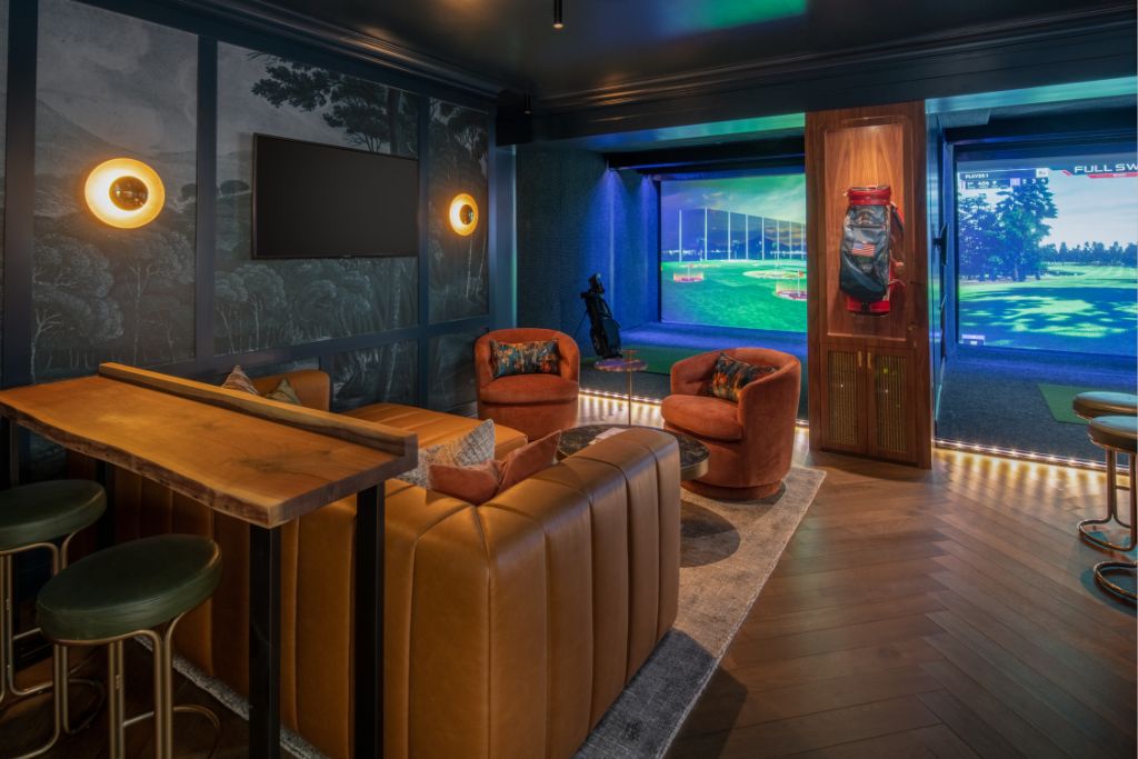 A modern home lounge with brown leather seating, a high table with stools, a wall-mounted TV, and a golf simulator setup in the corner, illuminated by warm lighting and featuring dark walls and wood flooring.